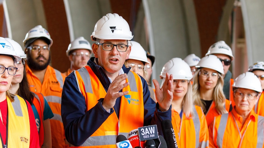 Daniel Andrews stands in hard hat and high-vis, holding hands out as he speaks at a press conference.