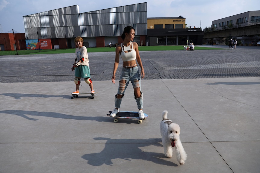 Twom women ride skateboards and a white fluffy dog runs. 