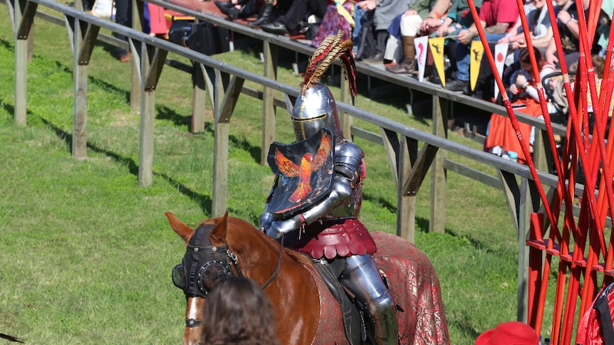 A jouster sits fully armoured on a horse with the crowds sitting on the stands in the background.