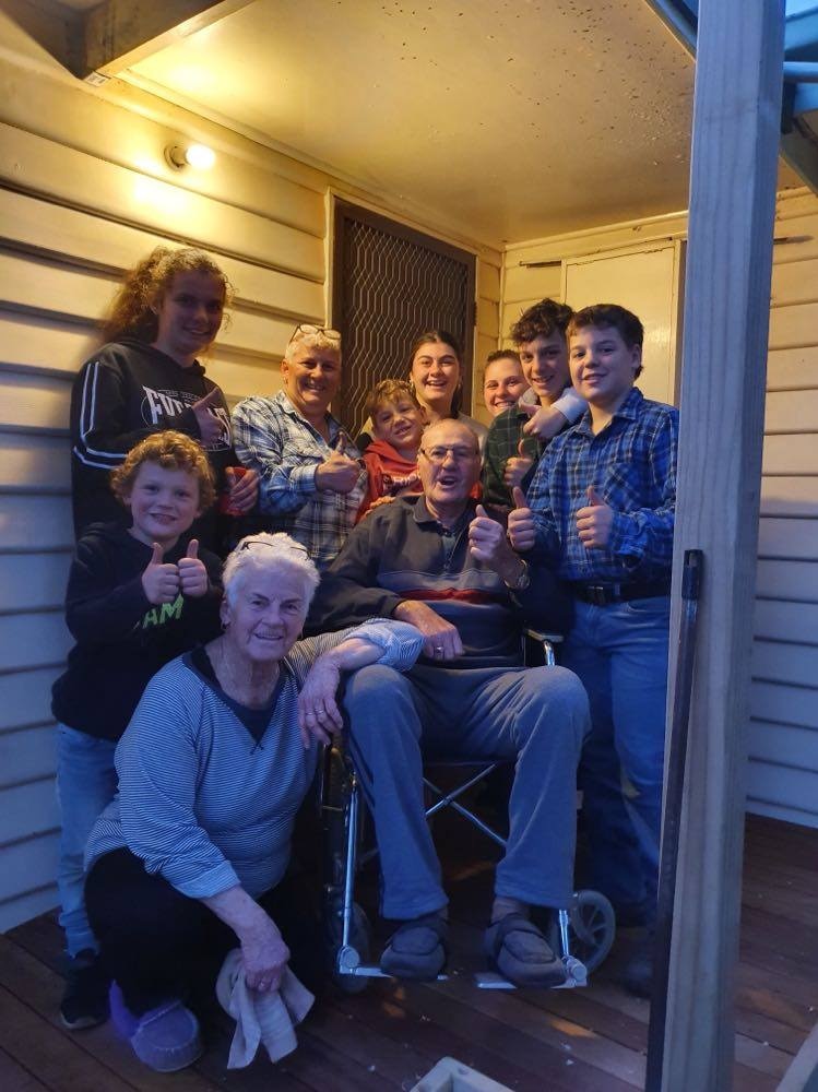 man in chair on front deck surrounded by family holding thumbs up