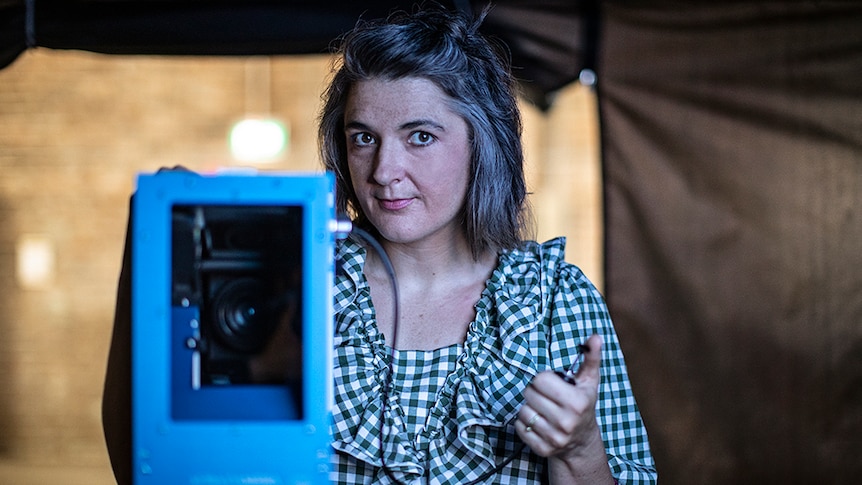 A woman with short dark and grey hair in white and dark green checked shirt stands in dark tent with camera equipment.
