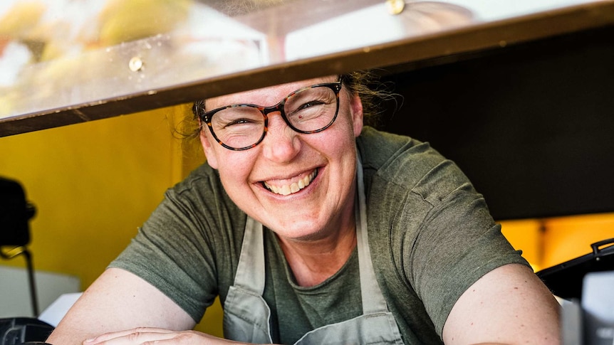 A woman wearing glasses and an apron beams a large smile leaning out a till window.