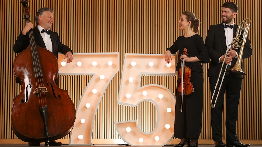 three musicians holding various instruments and dressed in black standing next to giant lights in the shape of the number '75'