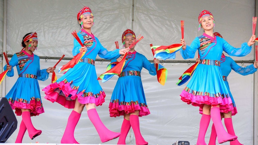Women dressed in bright blue and pink dresses perform on stage. They wear red headdresses and hold colourful decorations.