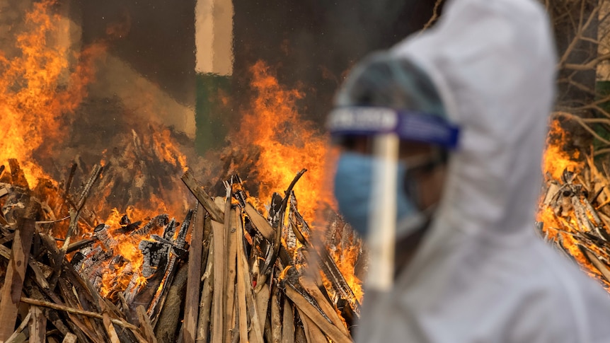 A man wearing white scrubs and a mask and shield walks by a fire.