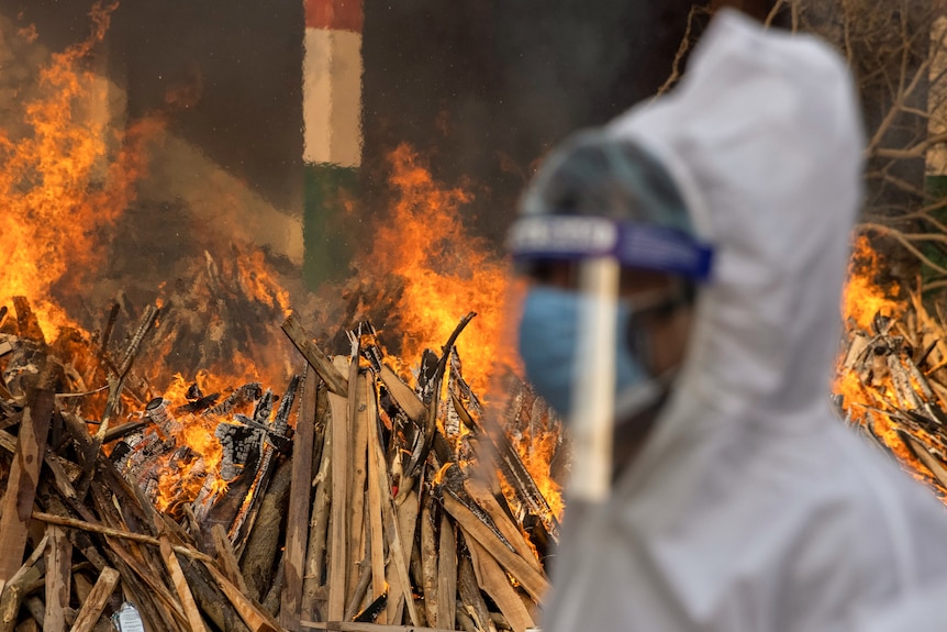 A man wearing white scrubs and a mask and shield walks by a fire.