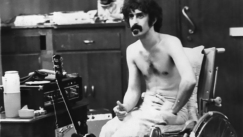A black and white photo of Frank Zappa sitting next to a guitar.