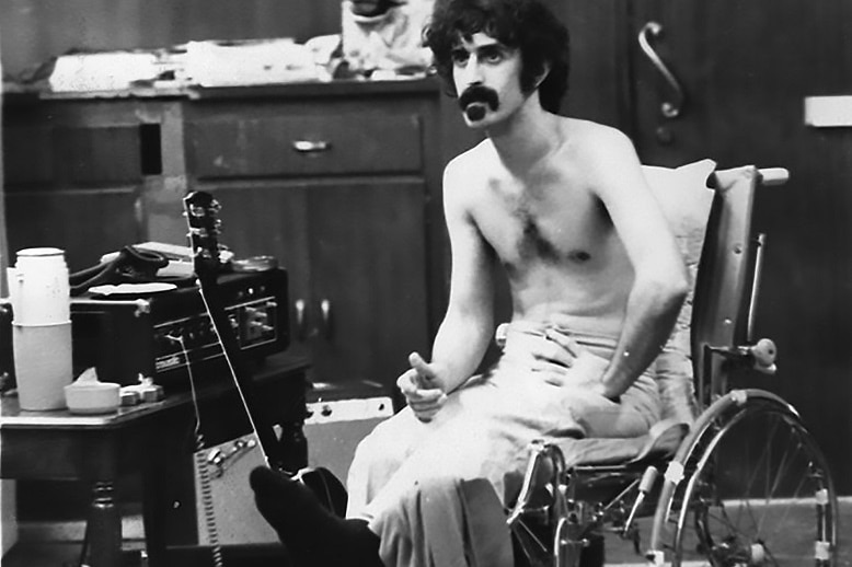 A black and white photo of Frank Zappa sitting next to a guitar.