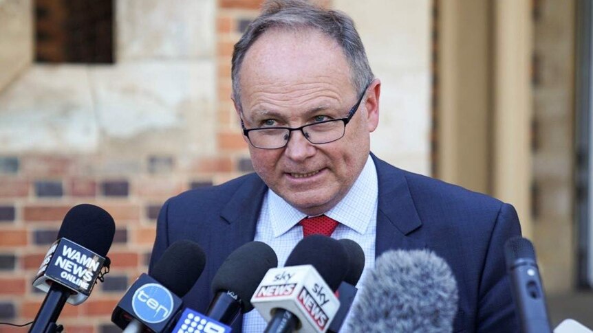 David Templeman wearing a suit and red tie speaks at a press conference in front of a bank of microphones.