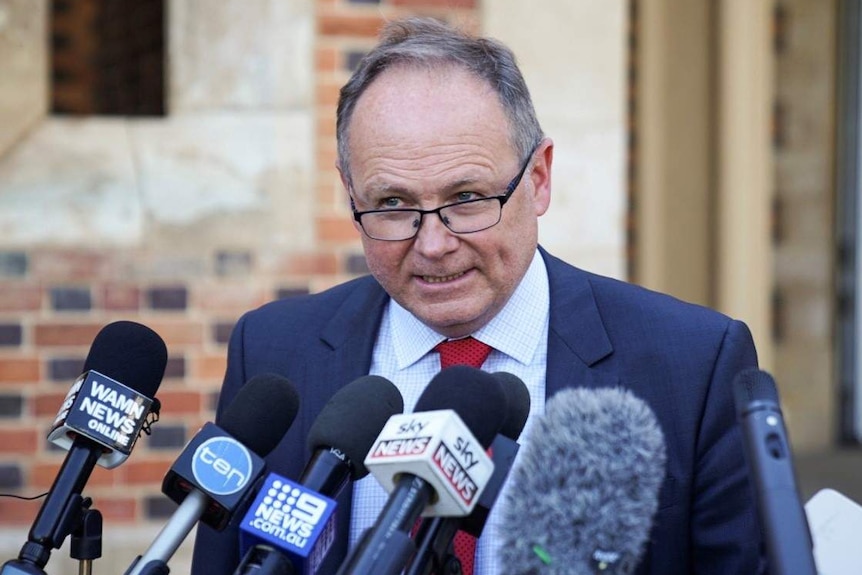 David Templeman wearing a suit and red tie speaks at a press conference in front of a bank of microphones.