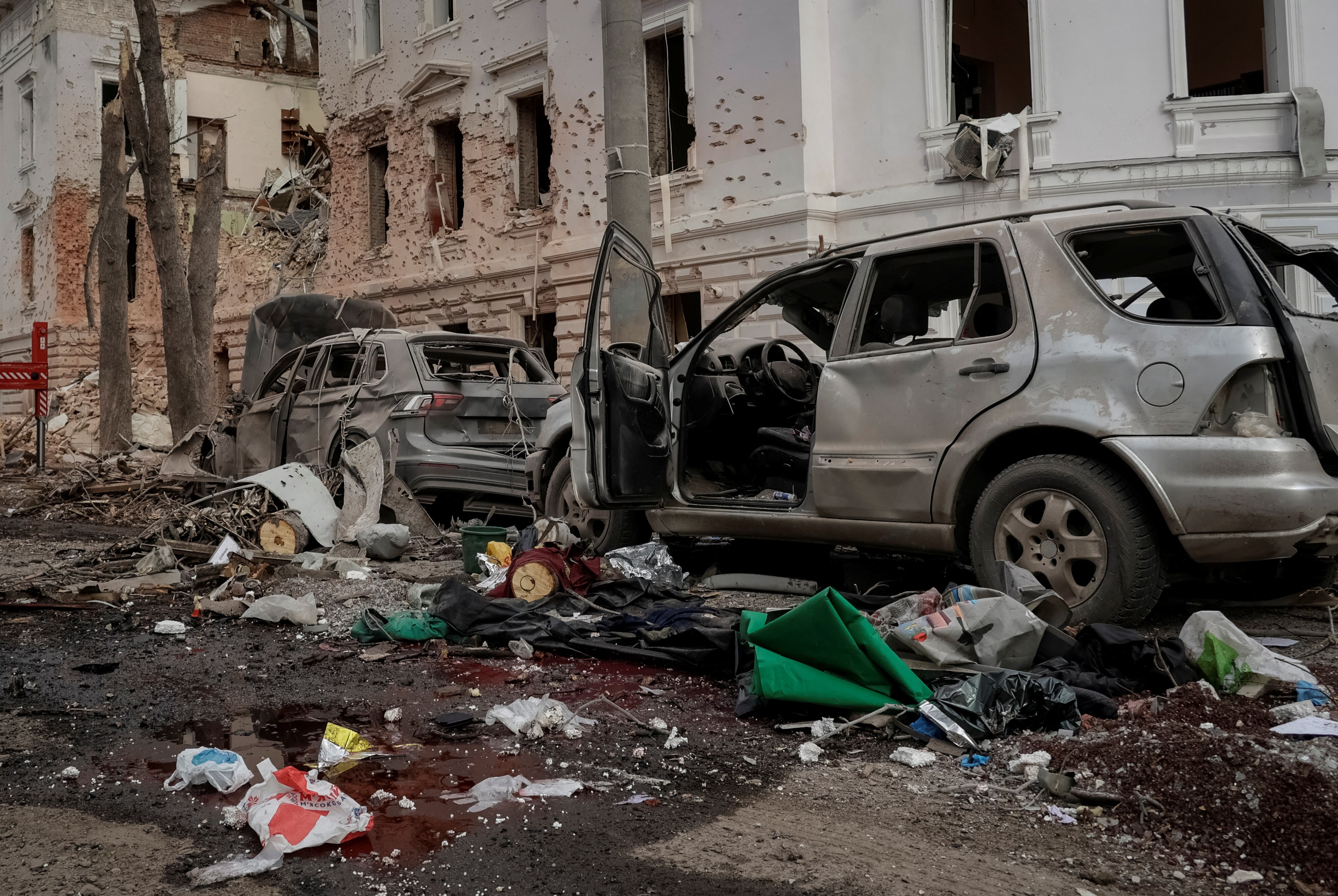 Two damaged cars alongside a road cluttered by debris and blood and buildings damaged by missiles
