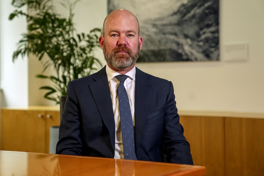 Homme chauve avec barbe portant costume bleu et cravate assis dans la salle de bureau en face de la table en bois