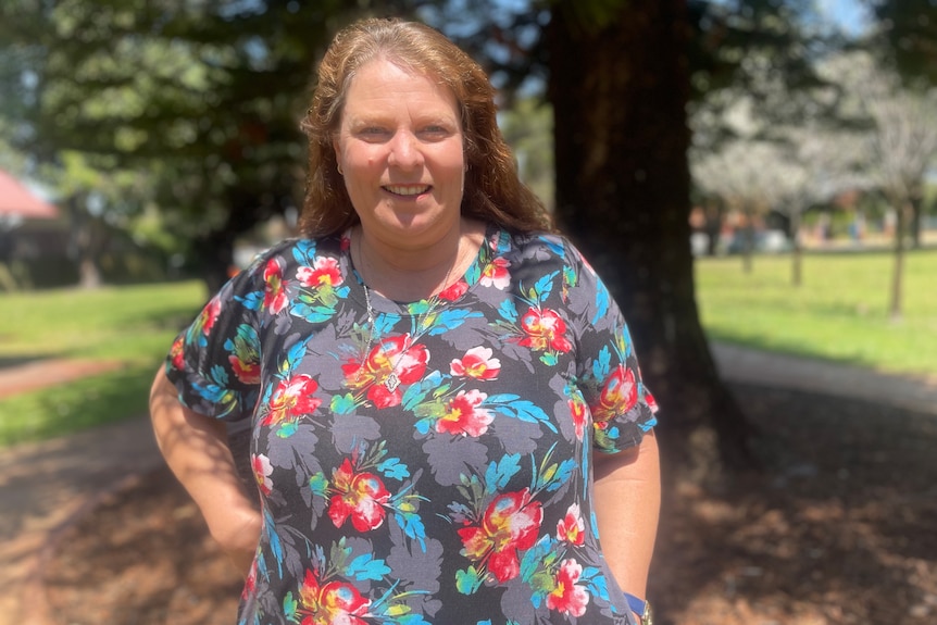 A woman stands outside in a park. There is a tree in the background.