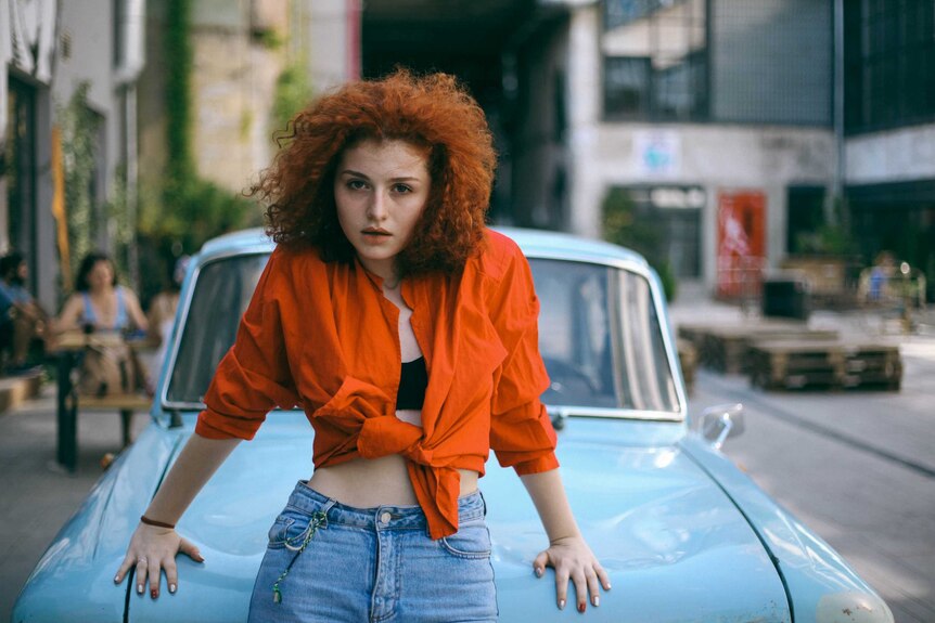 Woman with red, curly hair leans on a car in orange crop top and jeans
