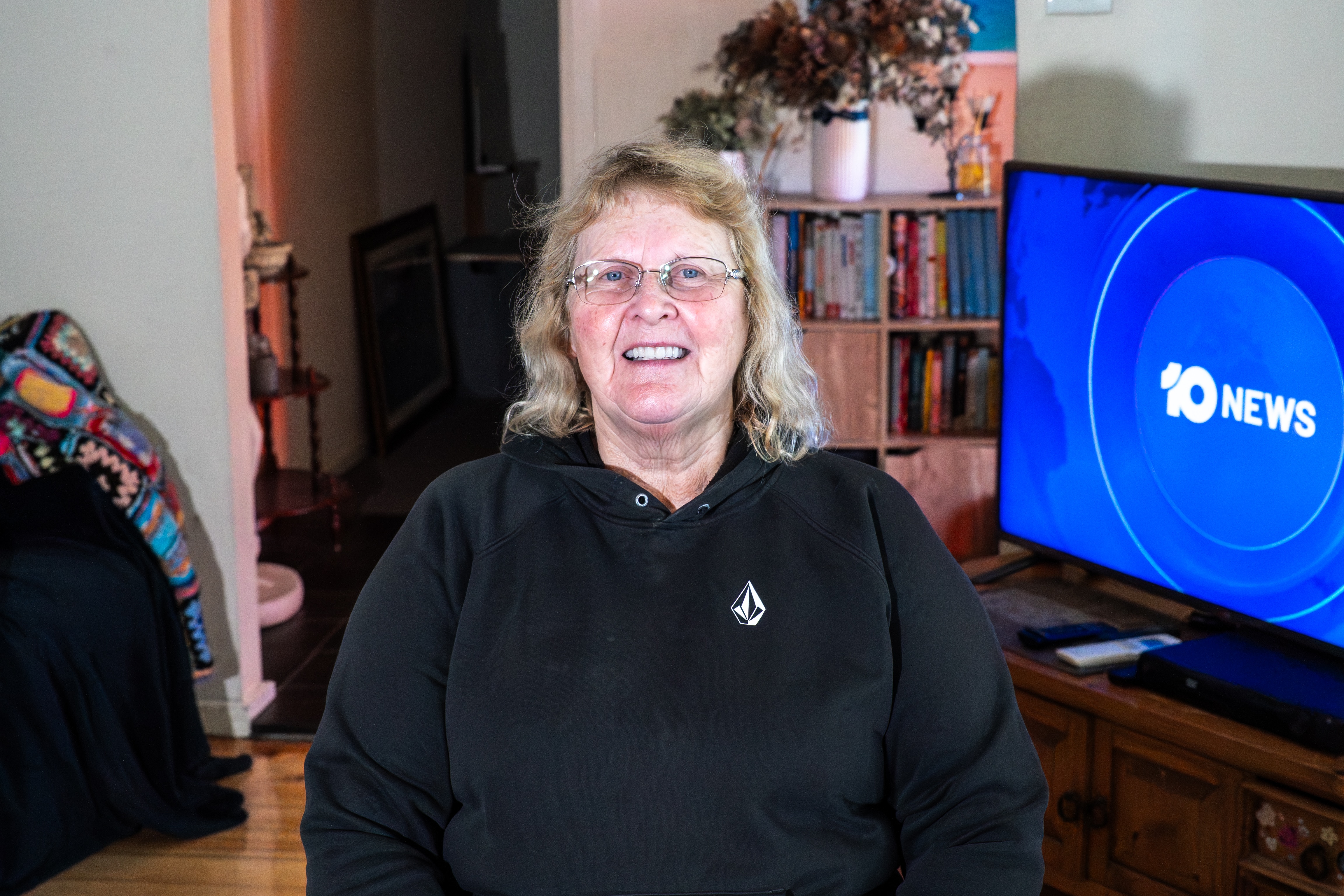 A woman sitting in a chair in her home, with a TV showing 10 News behind her