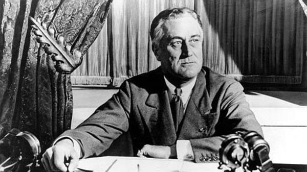 A black and white photograph of former US President, Franklin Delano Roosevelt sitting at his desk. 