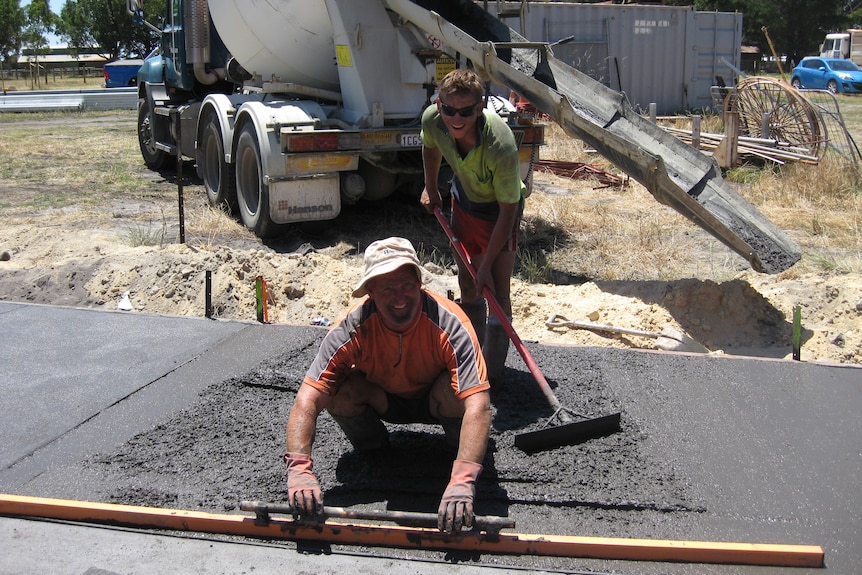 a man levelling concreting with a wooden plank , with a younger person working behind and a concrete truck in the background