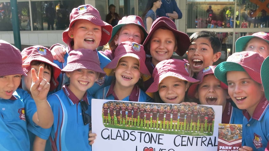 A group of school children wearing hats hold a sign welcoming the Queensland State of Origin team.