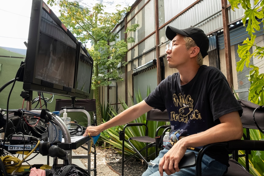 A behind the scenes pic of Lee Sung Jin outside on set sitting in a director's chair in front of a screen and equipment
