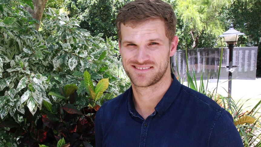 Young white man stands in front of greenery.