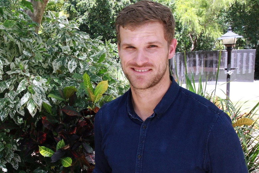 Young white man stands in front of greenery.