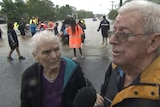 Elderly man and woman speaking into a microphone in front of floodwaters and rescue boats.