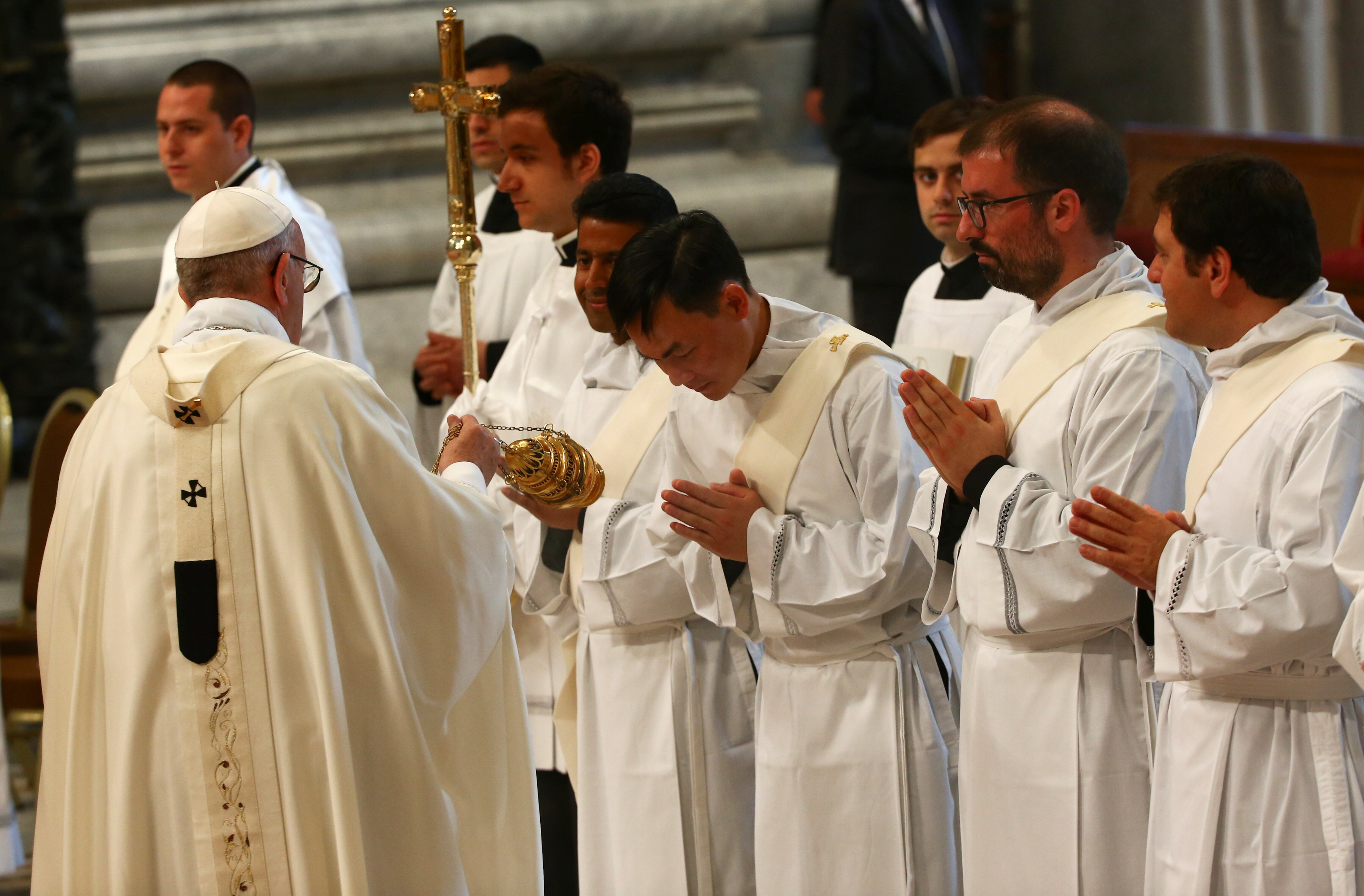 Pope Francis is seen in white robes, ordaining a line of priests standing in front of him with heads bowed.