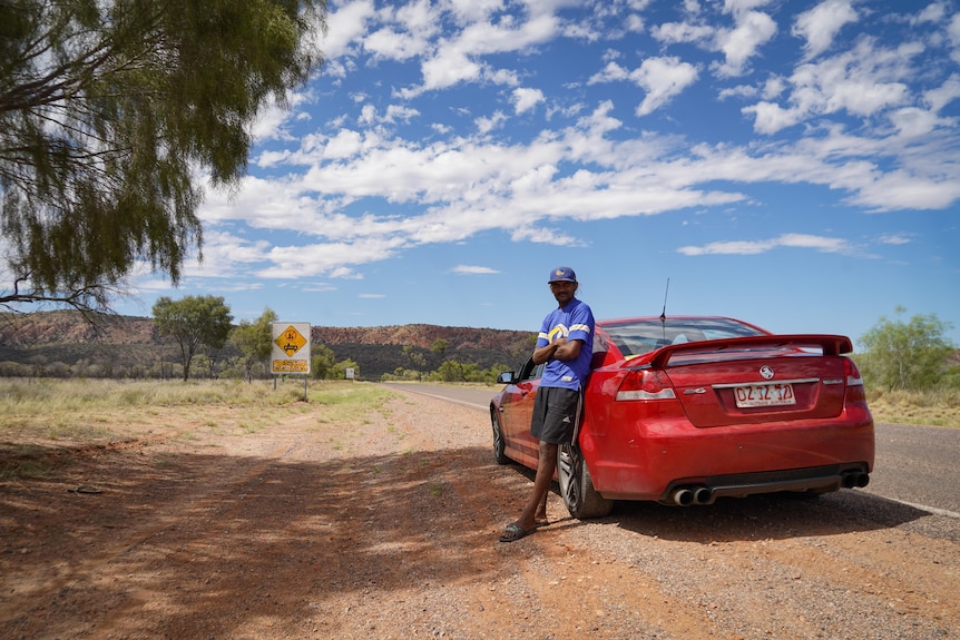 Man leans against red car with desert hills in the background 