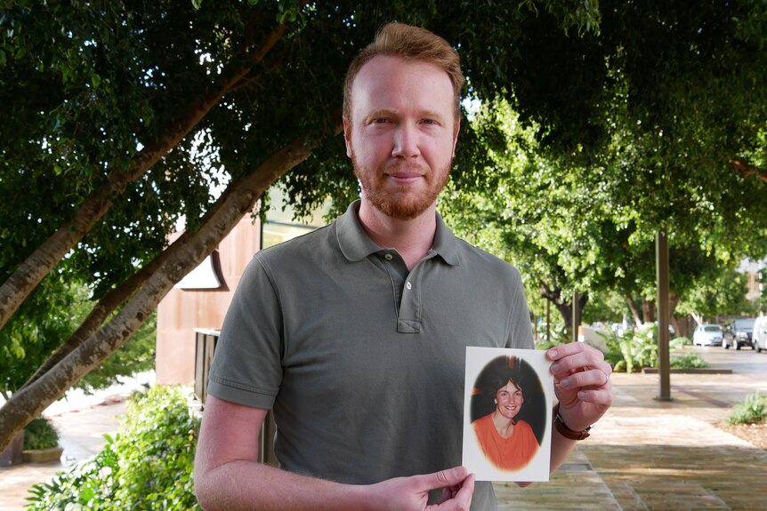 A man with red hair and a beard stands on a footpath, holding a photo of a woman.
