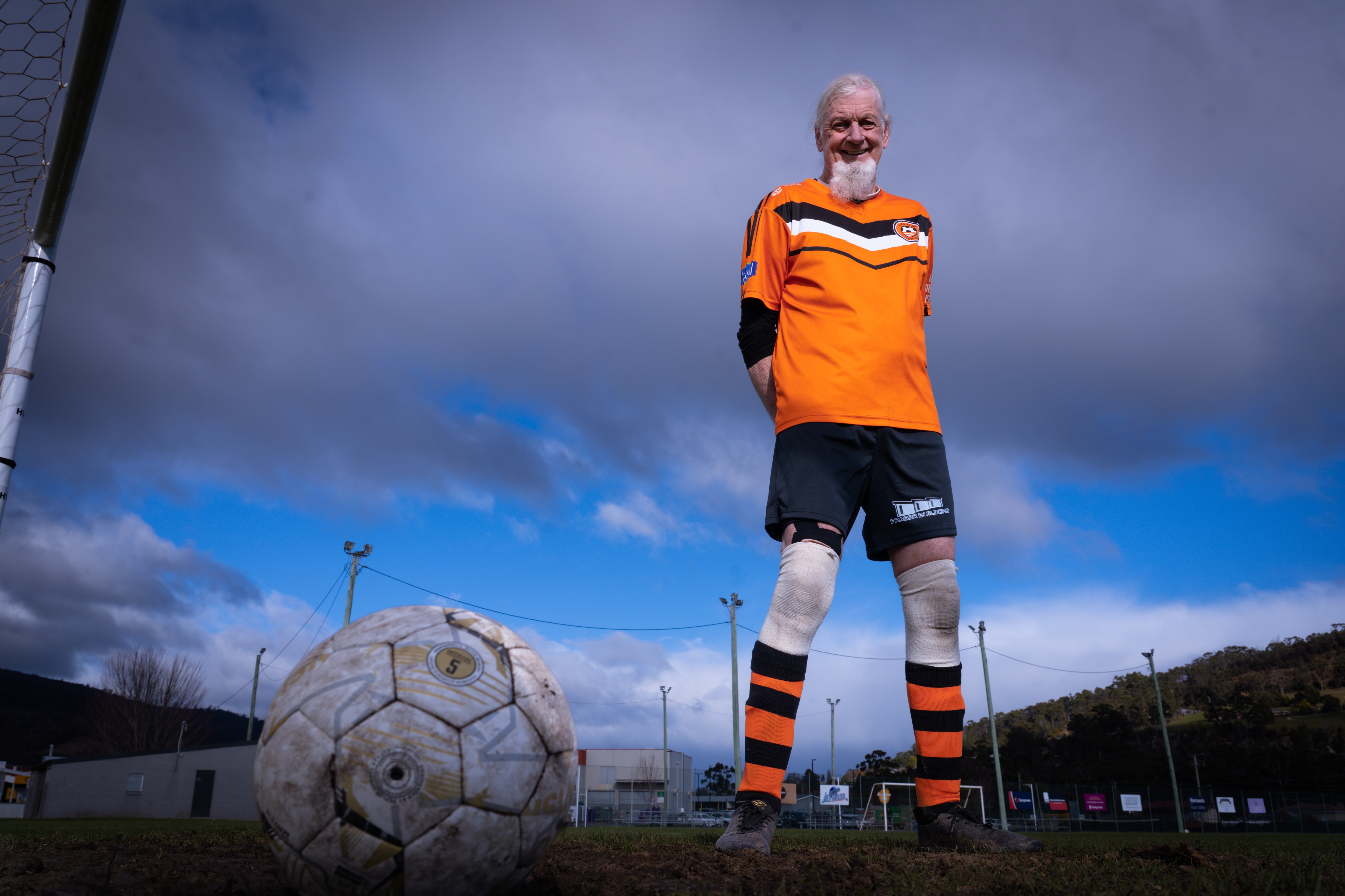 A man with white hair and a long white beard wearing orange soccer colours on a soccer field.