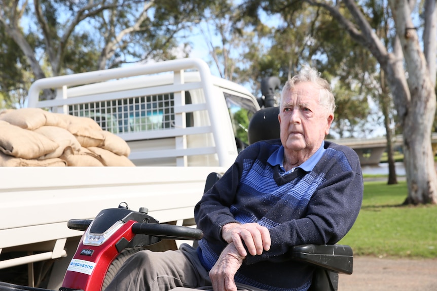 A man sits on a red motorised scooter beside a ute with sandbags in the tray.