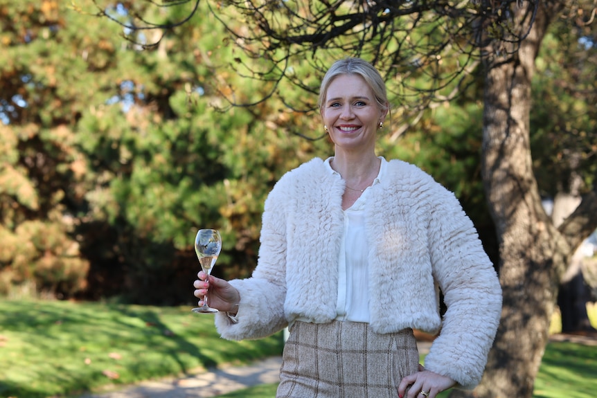A smiling blonde woman holds a glass of wine in a pleasant outdoor setting.
