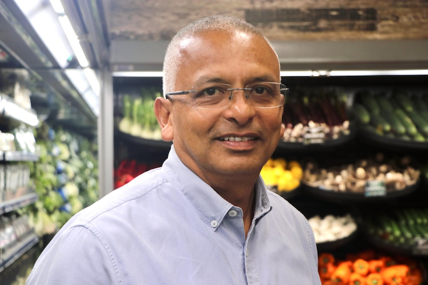 A man with glasses and very short hair stands in the fresh food aisle of a grocery store