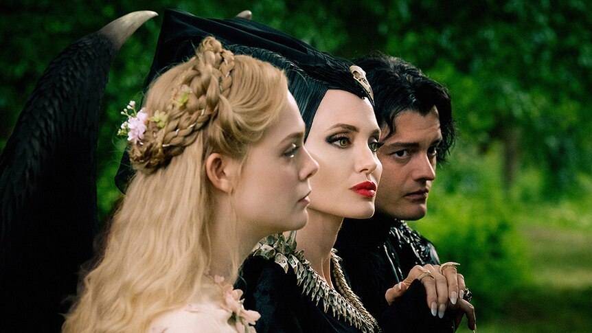 Elle Fanning, Angelina Jolie and Sam Riley stand side by side look in outdoor lush forest setting.