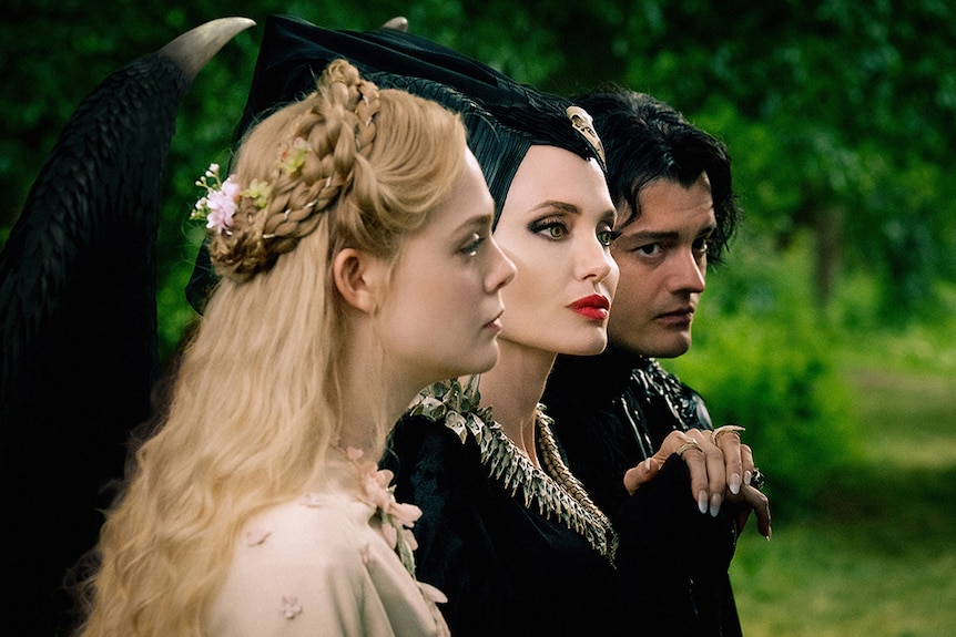 Elle Fanning, Angelina Jolie and Sam Riley stand side by side look in outdoor lush forest setting.