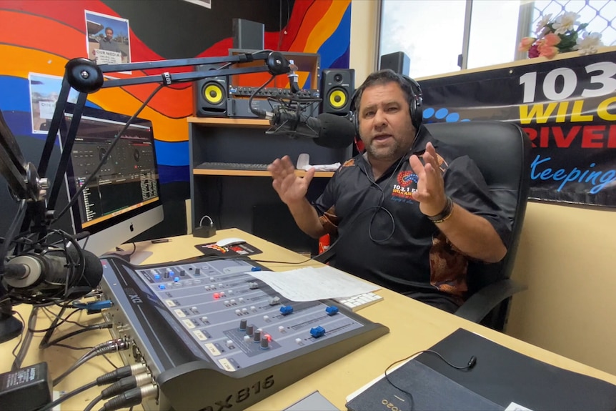 A man in a radio studio wearing headphones talking in to a microphone.