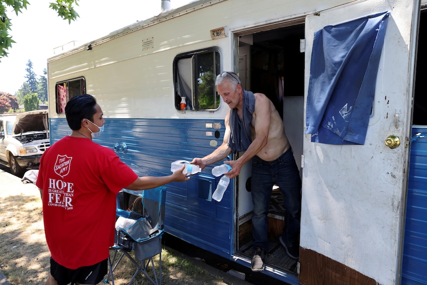 A man in a Salvation Army t-shirt hands water bottles to another man wearing no shirt in a caravan.