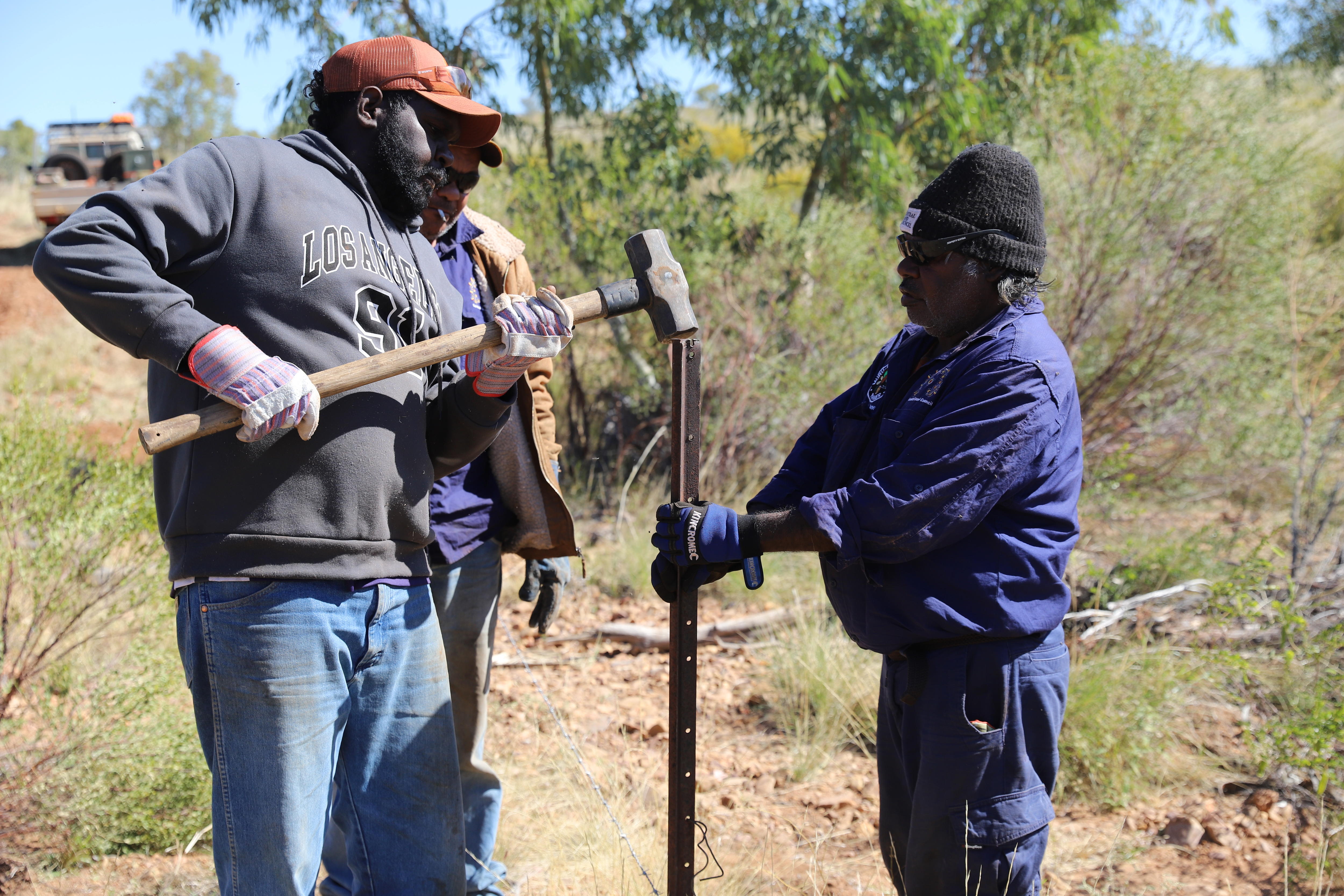 Three Aboriginal men huddle around a fence post, one uses a sledge hammer to bang it into the ground.
