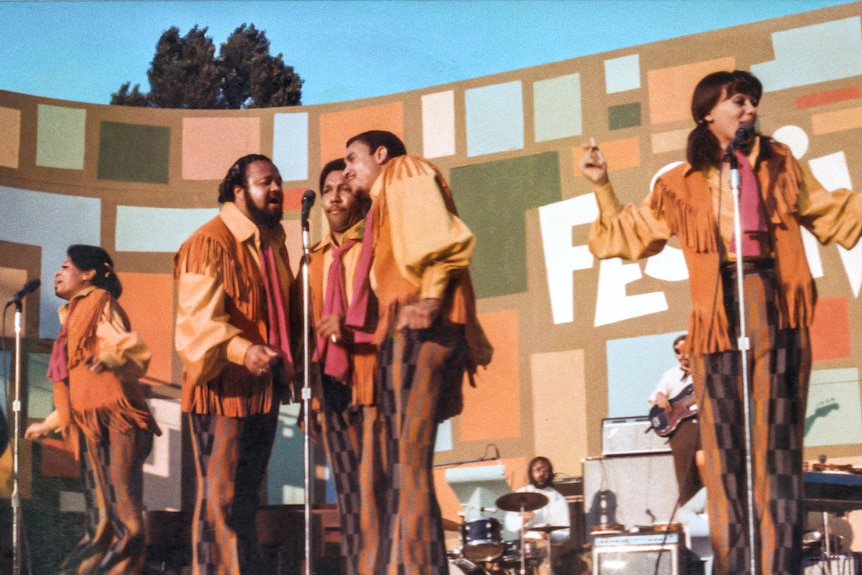 The band 5th Dimension singing on an outdoor stage in orange and yellow tassled 70s outfits, snapping their fingers to music.