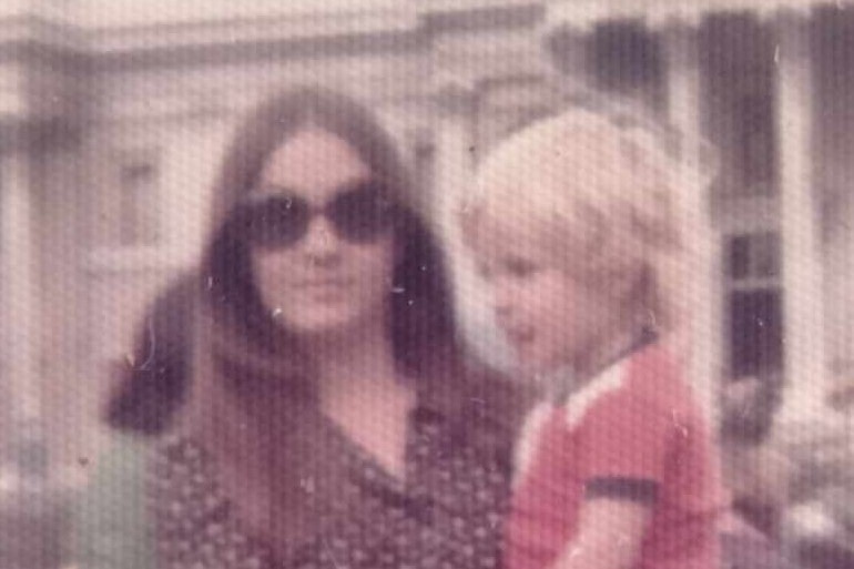 Old coloured photo of a woman in large sunglasses holding a small blonde-haired boy in her arms.