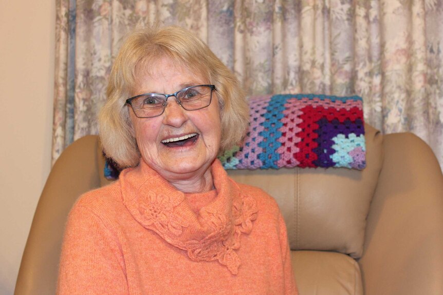 Joyce Watts, a dementia carer, smiles at camera. She wears glasses and an orange knit jumper. Sits on couch.