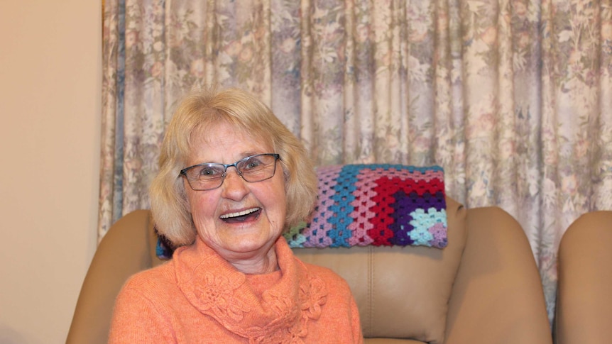 Joyce Watts, a dementia carer, smiles at camera. She wears glasses and an orange knit jumper. Sits on couch.