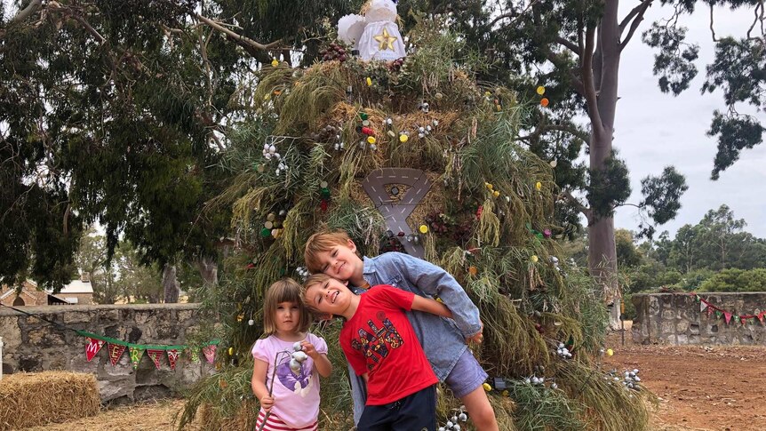 Three children frolic in front of a large decorated Christmas tree by the roadside.