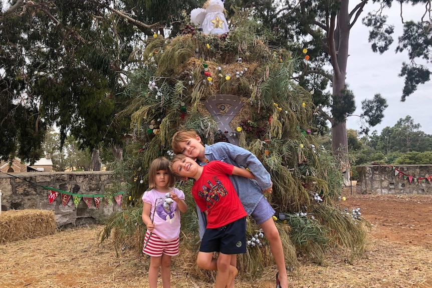 Three children frolic in front of a large decorated Christmas tree by the roadside.