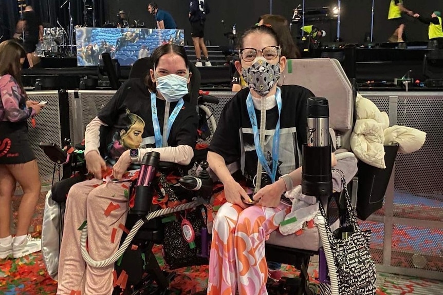 Two women wear ventilators and masks while sitting in wheelchairs at a concert