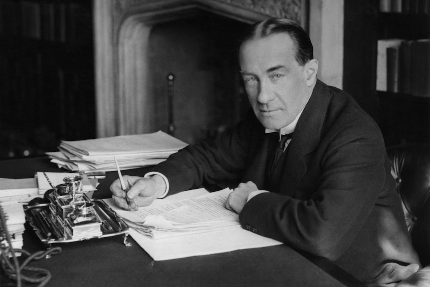 Stanley Baldwin is pictured sitting at his desk in a suit, writing on a piece of paper.