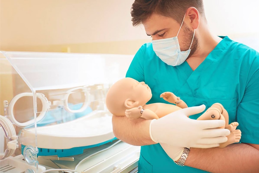 Man in blue scrubs nursing a Caucasian mannequin baby in front of an incubator.