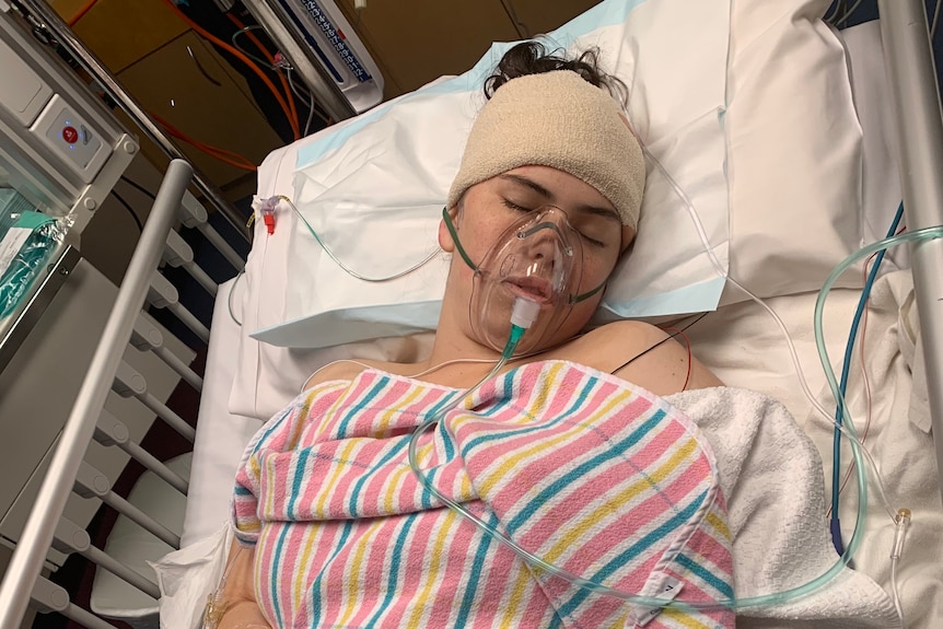 A girl in a hospital bed with a bandage around her head and an oxygen mask on