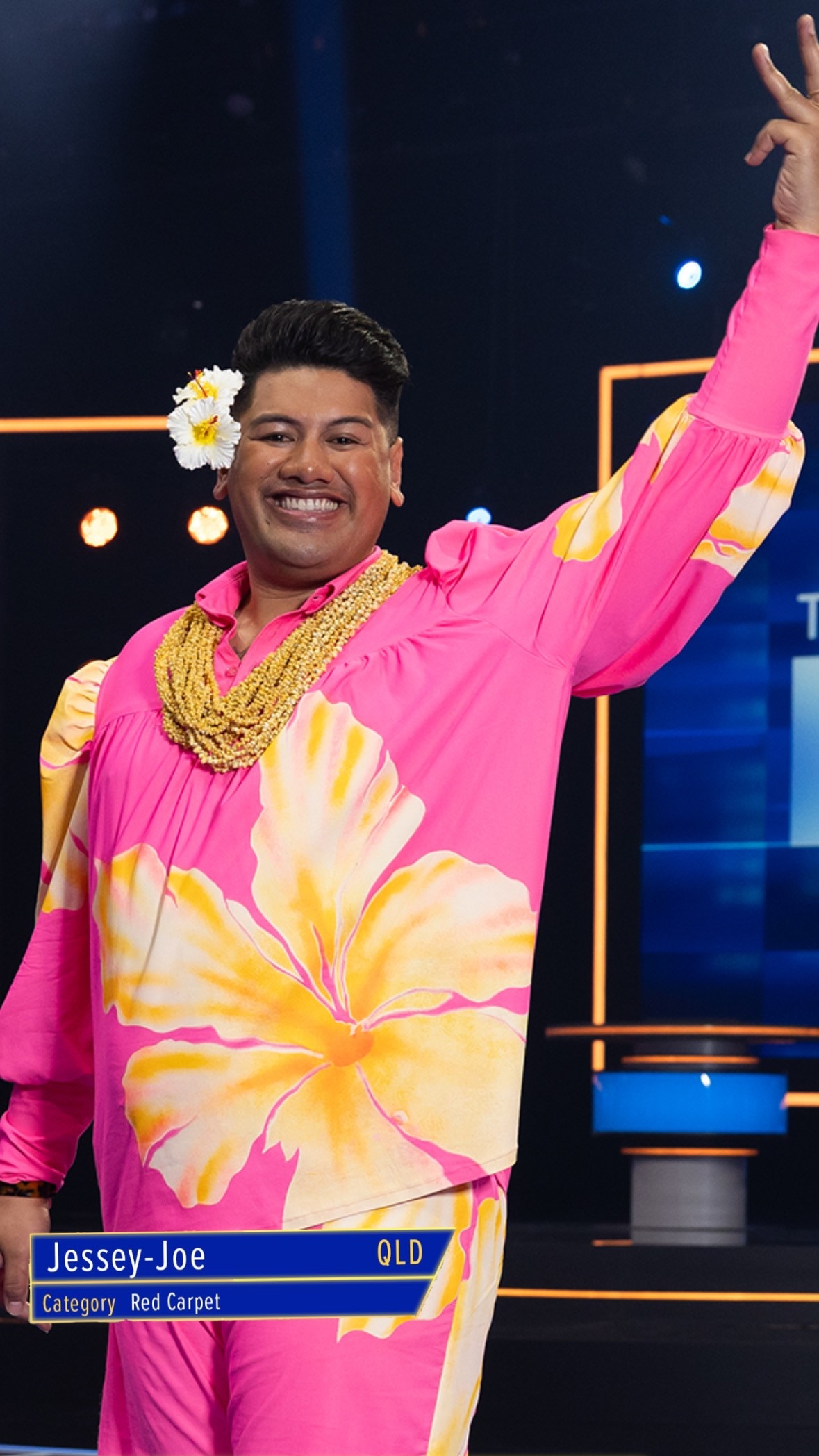 A man in a pink shirt and a flower in his ear waving.