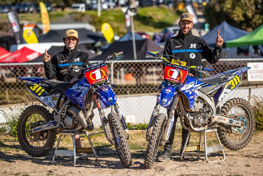 A man and a teenager holding up one finger standing in front of motorbikes at a motorcross competition